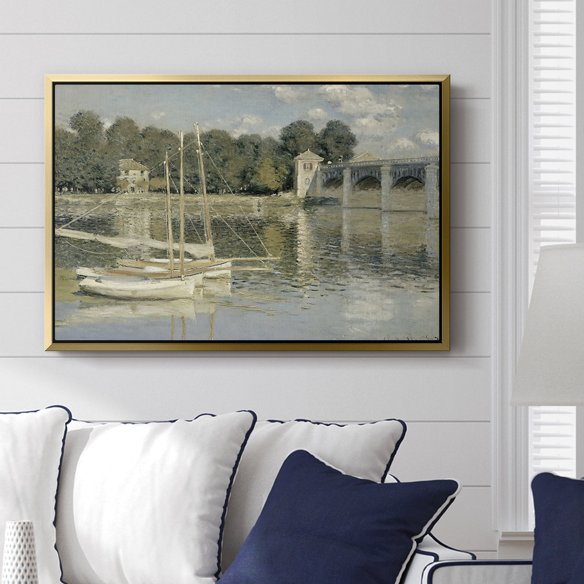 The Bridge at Argenteuil, 1874 by Claude Monet displayed above a sofa in a living room in a gold floater frame, featuring an impressionist river scene with boats passing beneath bridge.