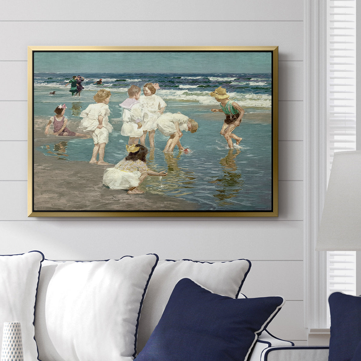 A Holiday, c. 1915 by Edward Henry Potthast displayed above a sofa in a living room in a gold floater frame, featuring a beach scene with figures bathing and standing in waves under bright sky.