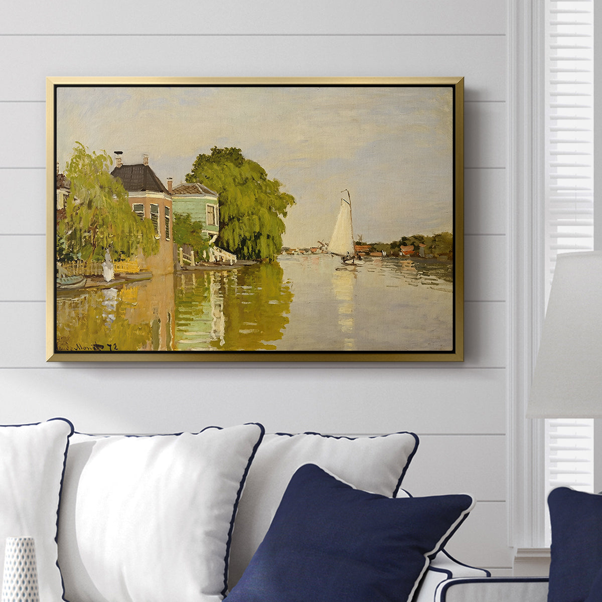 Houses on the Achterzaan, 1871 by Claude Monet displayed above a sofa in a living room in a gold floater frame, featuring riverside homes with trees and mirrored reflections in water.