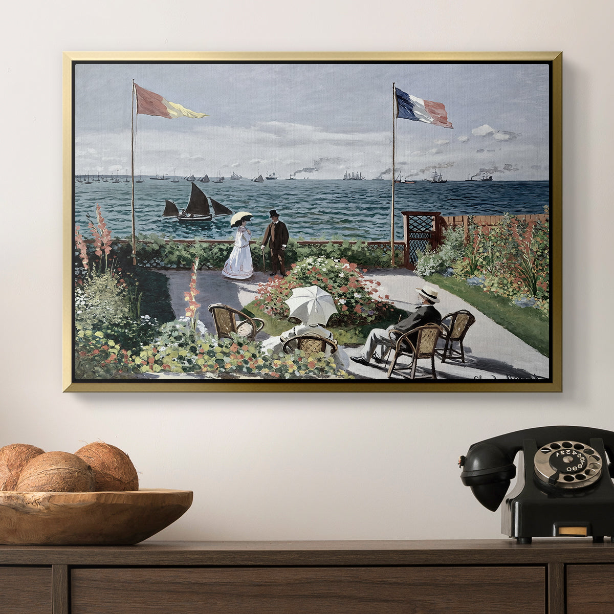 Garden at Sainte-Adresse, 1867 by Claude Monet in a gold floater framed canvas displayed above a console table in a hallway, featuring a landscaped terrace with figures and flags near shoreline.