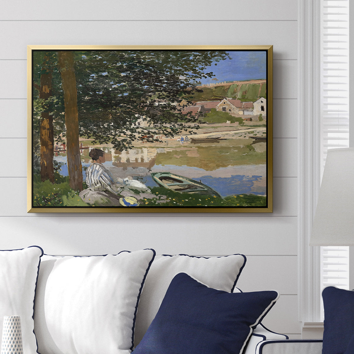 On the Bank of the Seine, Bennecourt, 1868 by Claude Monet displayed above a sofa in a living room in a gold floater frame, featuring a peaceful riverside scene with figures resting in grassy area by water.
