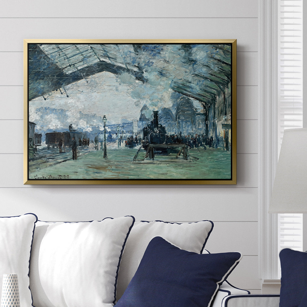 The Saint-Lazare Station, 1877 by Claude Monet displayed above a sofa in a living room in a gold floater frame, featuring an impressionist railway scene with locomotives and haze.