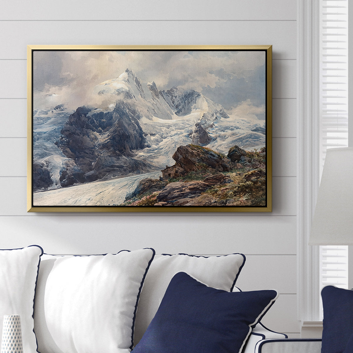 The Grossglockner, 1880 by Edward Theodore Compton displayed above a sofa in a living room in a gold floater frame, featuring a dramatic mountain summit with snow and rugged slopes.