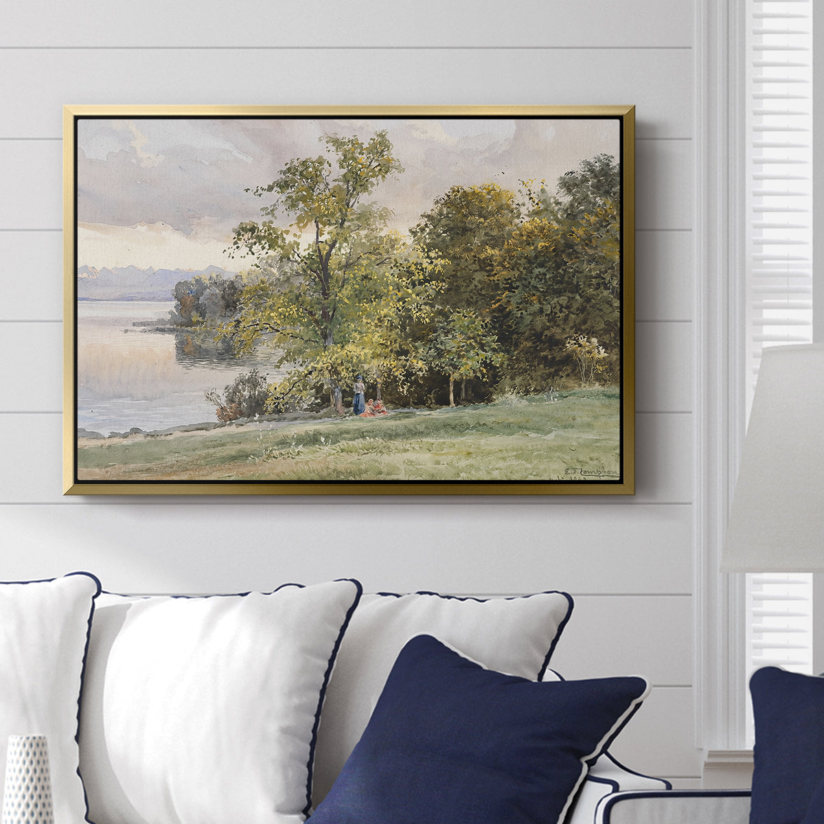 Landscape at Lake Starnberg, 1890 by Edward Theodore Compton displayed above a sofa in a living room in a gold floater frame, featuring a peaceful lake scene with reflections and surrounding greenery.