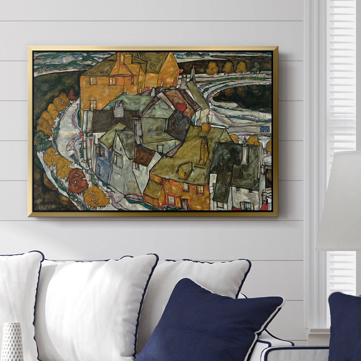 Crescent of Houses II (Island Town), 1915 by Egon Schiele displayed above a sofa in a living room in a gold floater frame, featuring a stylized townscape with clustered buildings and bold outlines.