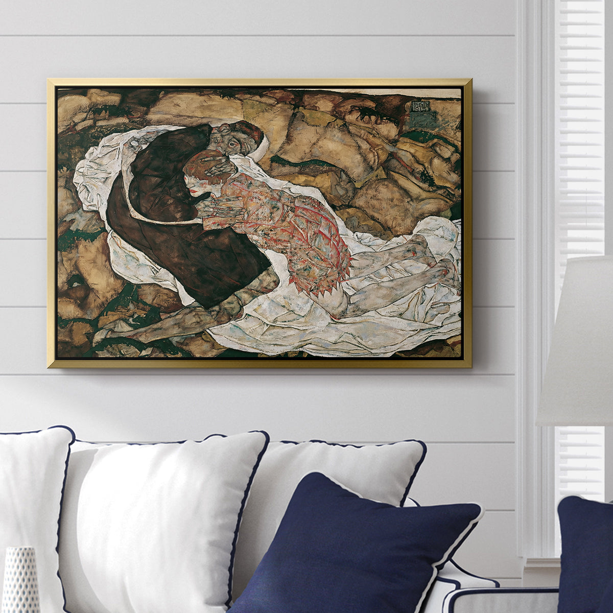 Death and the Maiden, 1915 by Egon Schiele displayed above a sofa in a living room in a gold floater frame, featuring a symbolic scene of mortality with two figures clinging together.