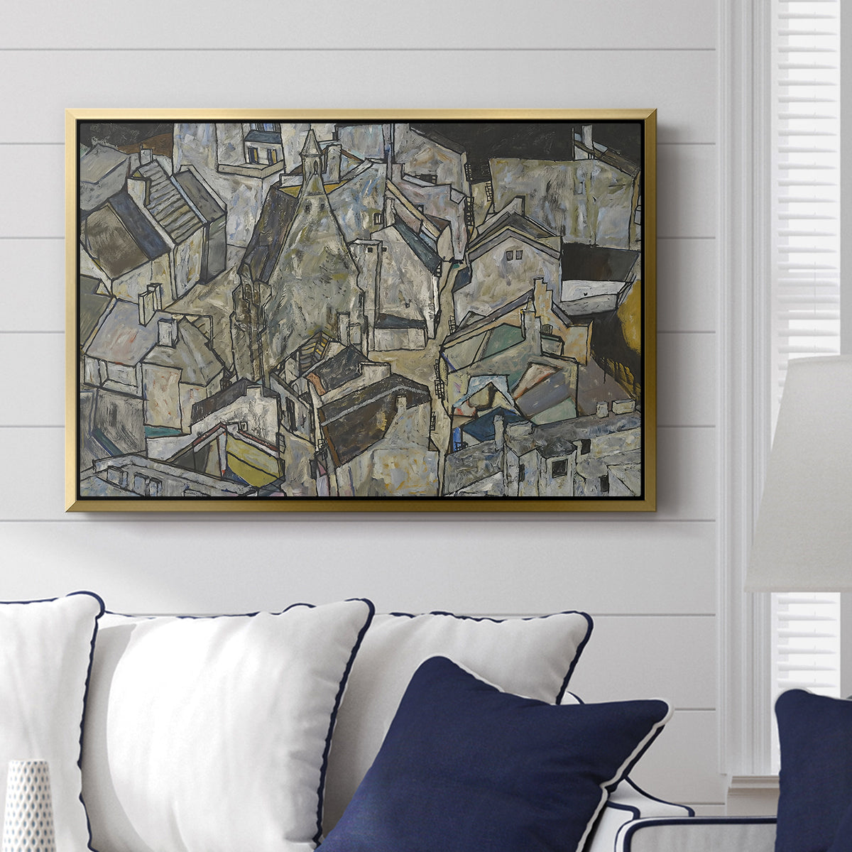 Mödling II, 1918 by Egon Schiele displayed above a sofa in a living room in a gold floater frame, featuring an expressionist cityscape with tightly packed buildings and bold lines.
