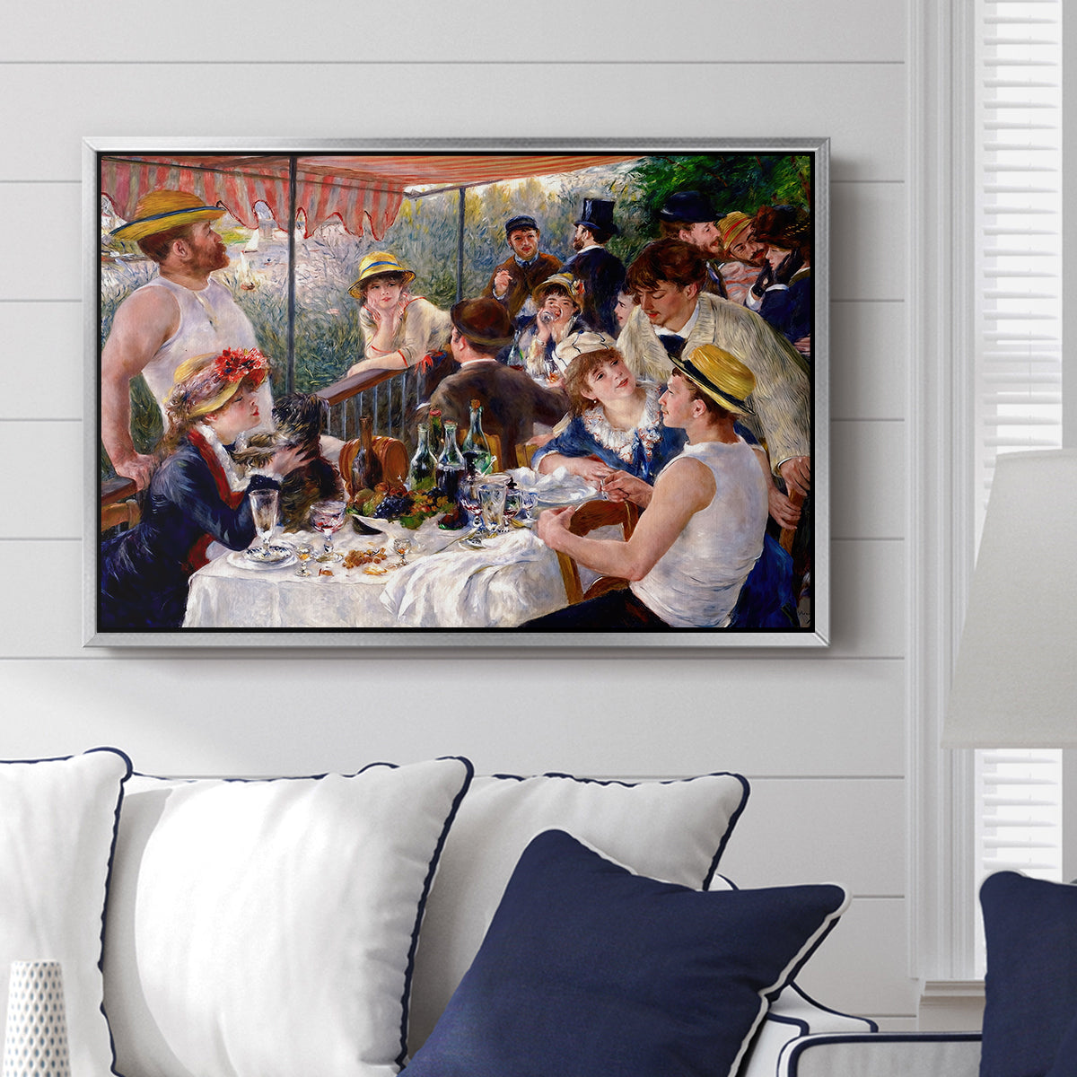 Luncheon of the Boating Party (Le Déjeuner des canotiers), 1880-1881 by Pierre-Auguste Renoir displayed above a sofa in a living room in a silver floater frame, featuring a lively gathering of men and women eating and socializing at outdoor table.