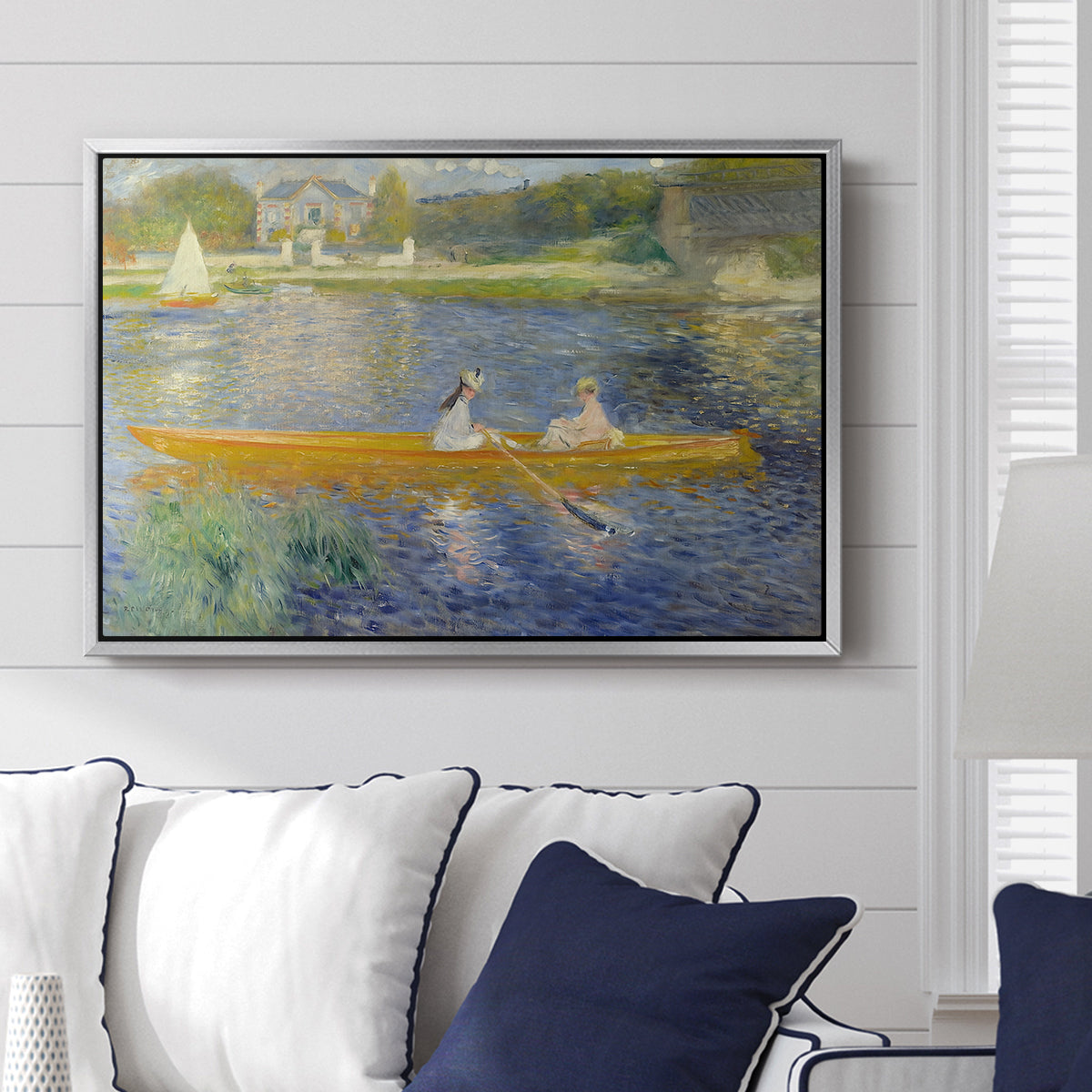 The Skiff (La Yole), 1875 by Pierre-Auguste Renoir displayed above a sofa in a living room in a silver floater frame, featuring an impressionist boating scene with people relaxing in a narrow skiff.