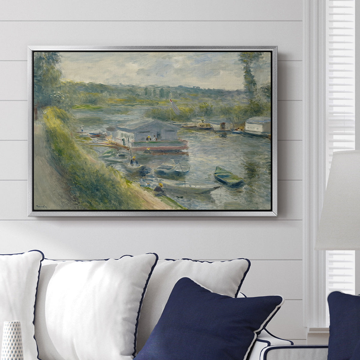 Wash-House Boat at Bas-Meudon, c. 1874 by Pierre-Auguste Renoir displayed above a sofa in a living room in a silver floater frame, featuring a riverside scene with moored boat used for washing clothes.