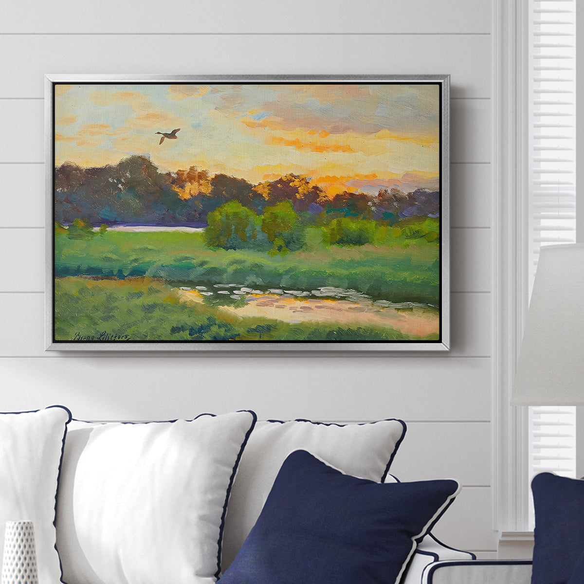 Flying Duck at Sunset, 1924 by Bruno Liljefors displayed above a sofa in a living room in a silver floater frame, featuring a lone duck soaring over a glowing marshland.