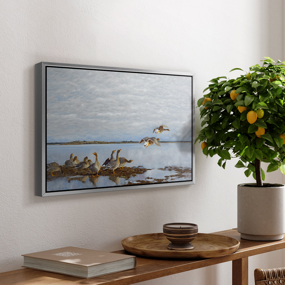 Greylag Geese (Grågäss), 1922 by Bruno Liljefors, presented in a silver floater framed canvas featuring a group of geese gathered near water in a natural wetland landscape, displayed above a hallway console table.