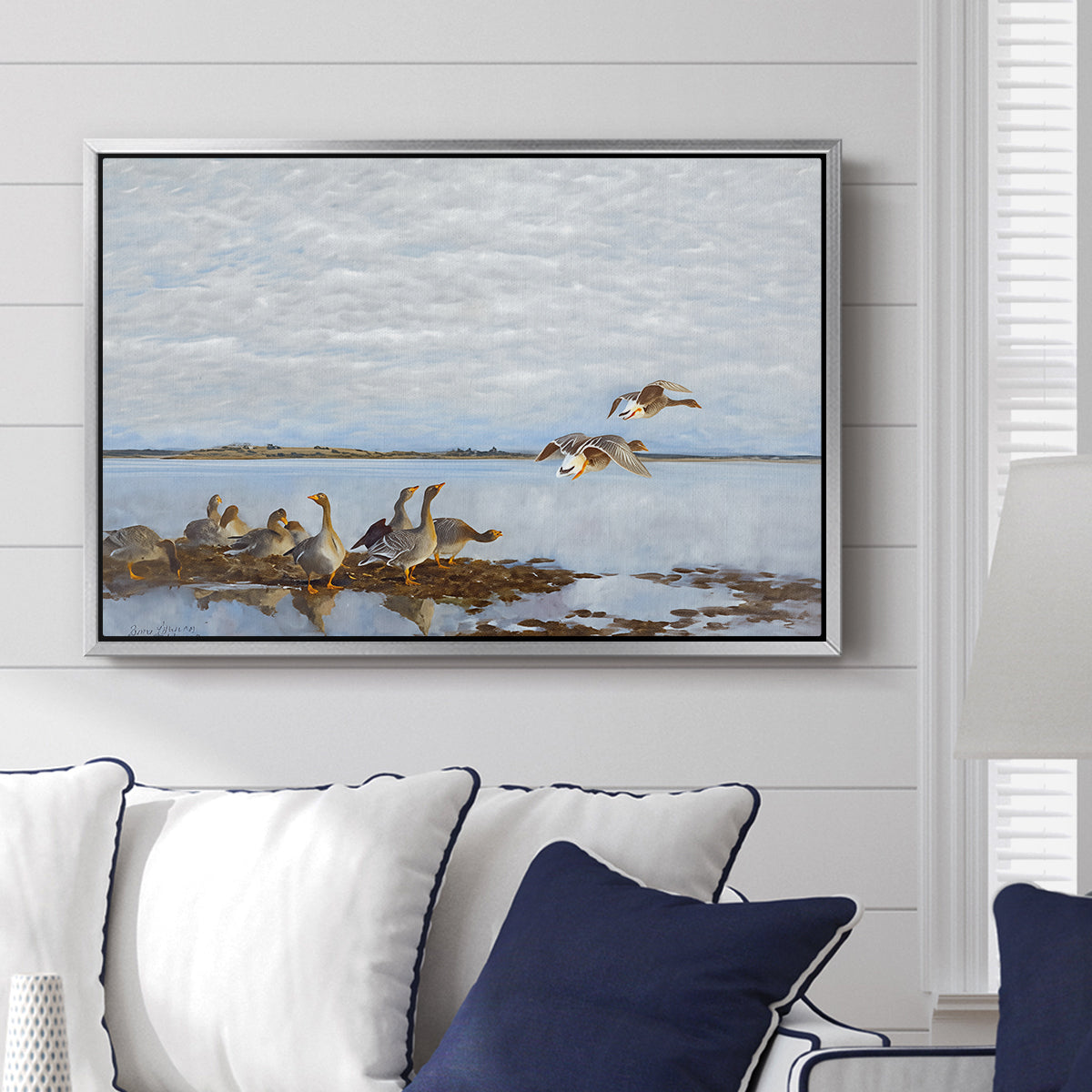 Greylag Geese (Grågäss), 1922 by Bruno Liljefors displayed above a sofa in a living room in a silver floater frame, featuring birds standing and resting along shoreline with soft grass and water.