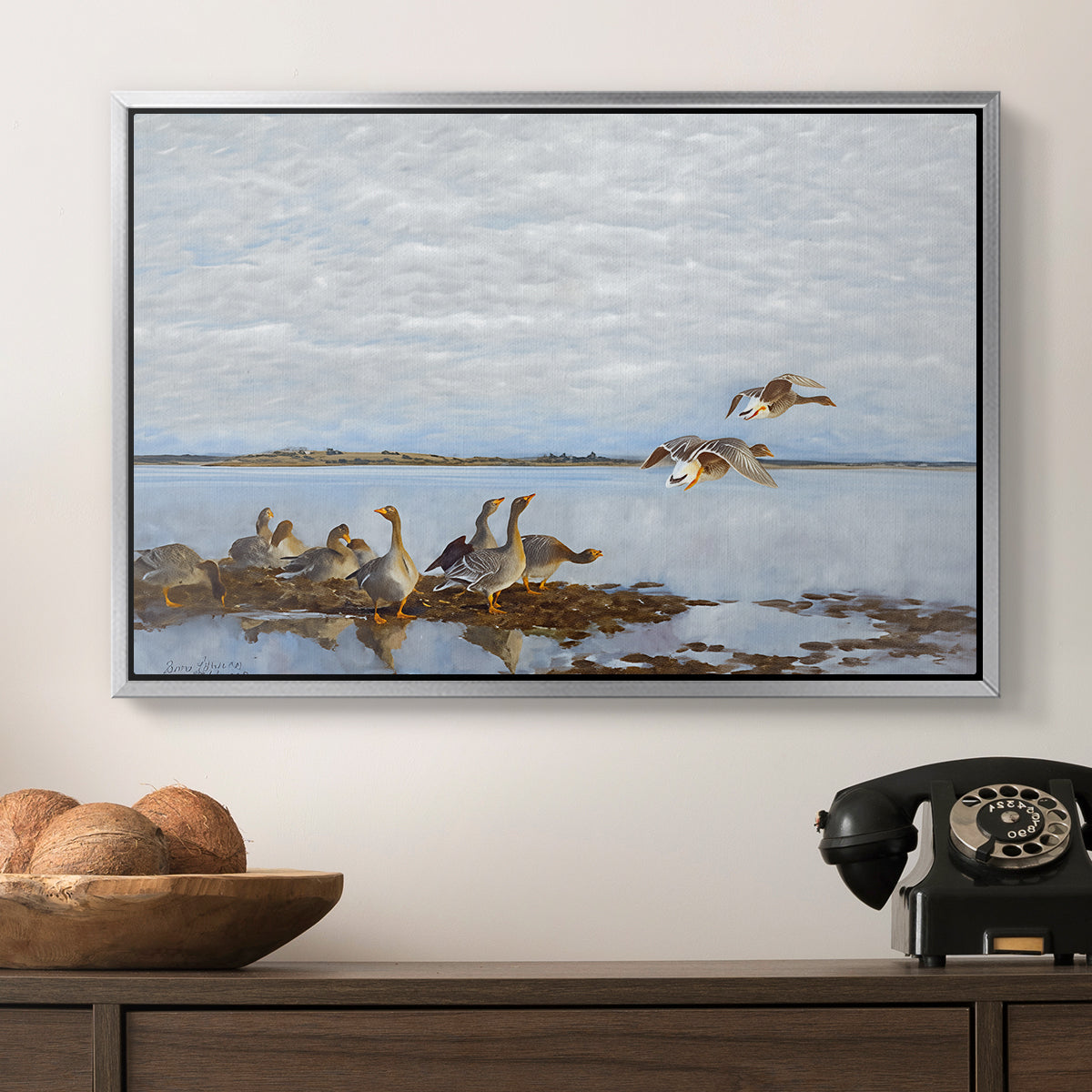 Greylag Geese (Grågäss), 1922 by Bruno Liljefors in a silver floater framed canvas displayed above a console table in a hallway, featuring a flock of geese positioned along marshy terrain.