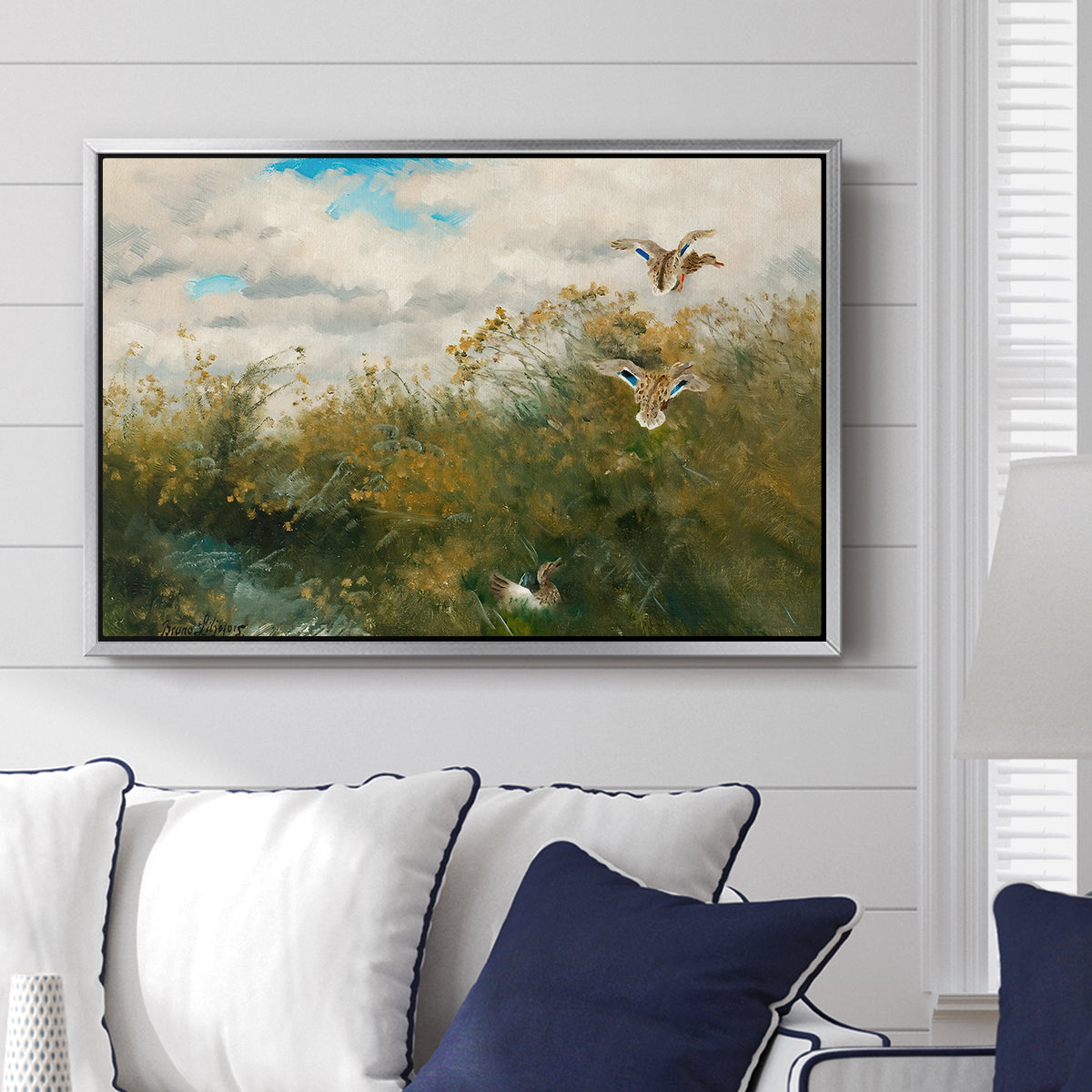 Wild Ducks (Grasänder), 1890 by Bruno Liljefors displayed above a sofa in a living room in a silver floater frame, featuring a wildlife scene of ducks walking through tall grass beside water.