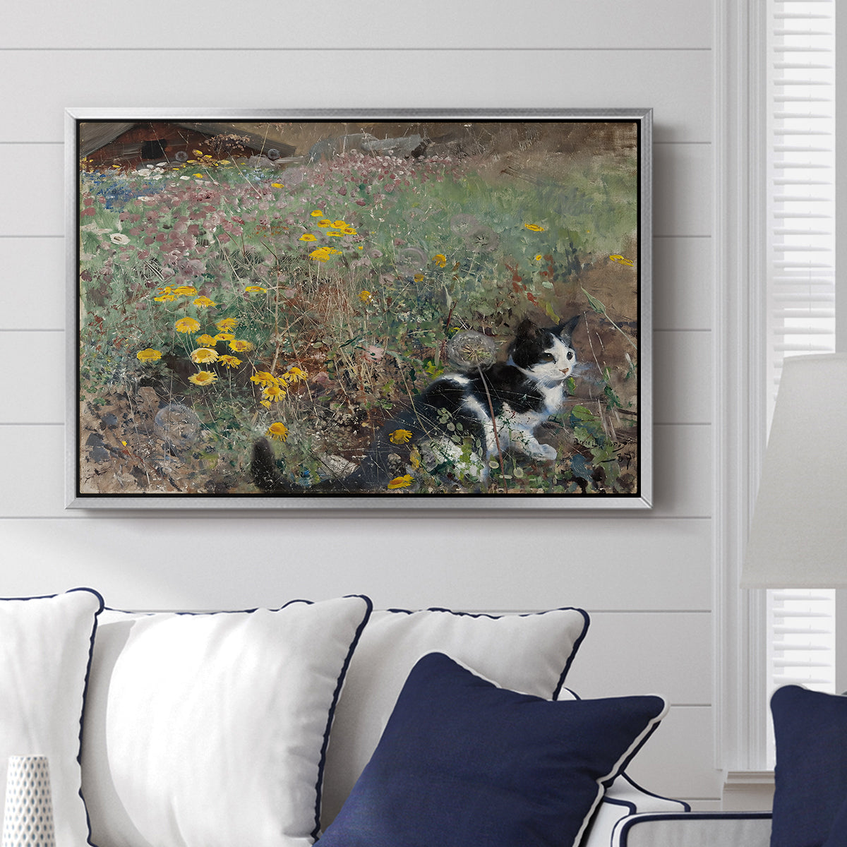 Cat on a Flowery Meadow, 1887 by Bruno Liljefors displayed above a sofa in a living room in a silver floater frame, featuring a small cat resting among colorful flowers in grassy field.