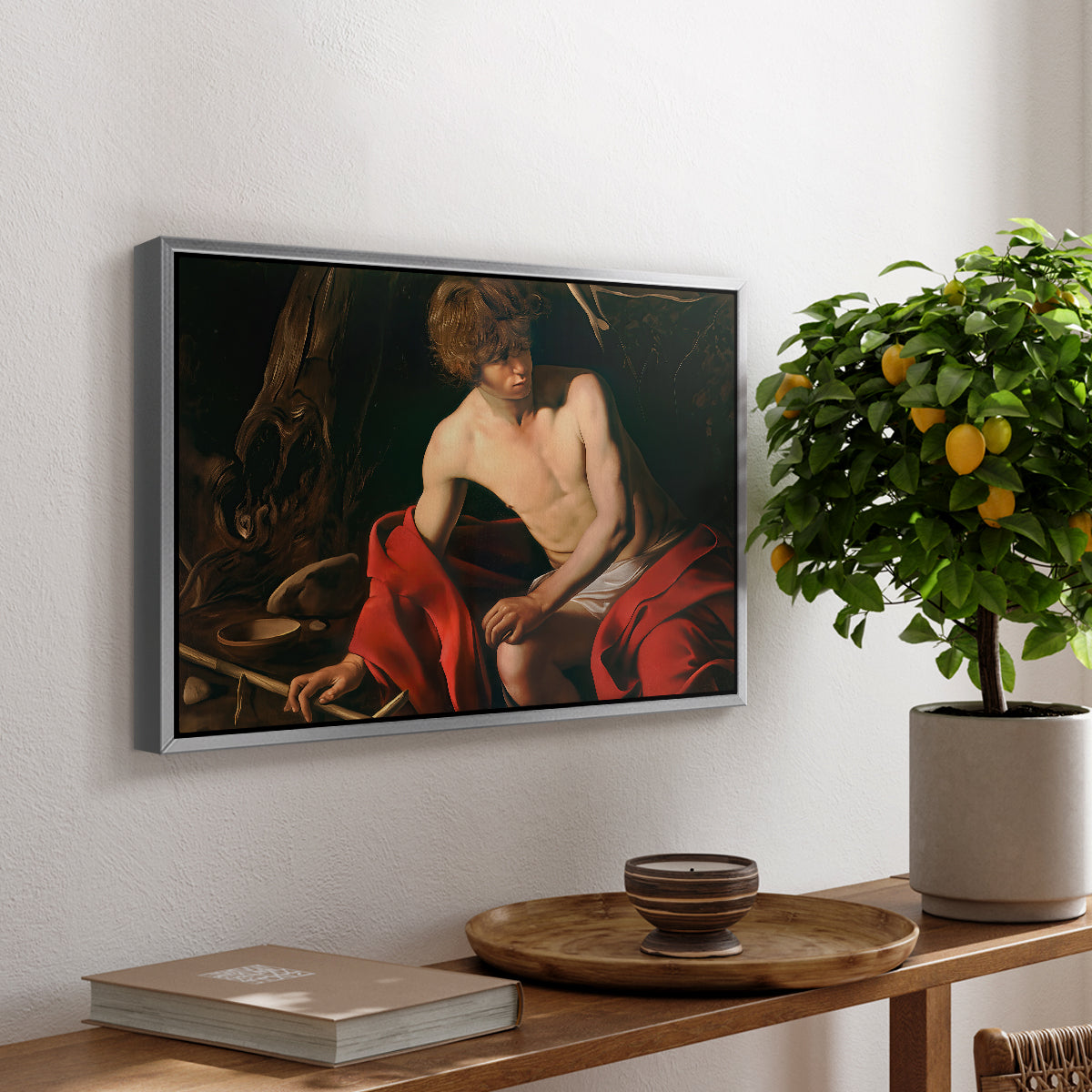 Saint John the Baptist in the Wilderness, c. 1603-1604 by Caravaggio, presented in a silver floater framed canvas featuring a young Saint John seated in wilderness with staff and red drapery, displayed above a hallway console table.