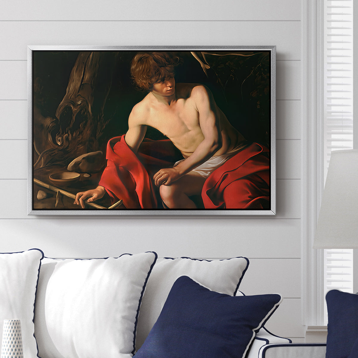 Saint John the Baptist in the Wilderness, c. 1603-1604 by Caravaggio displayed above a sofa in a living room in a silver floater frame, featuring a solitary figure of Saint John resting in natural setting with soft light.