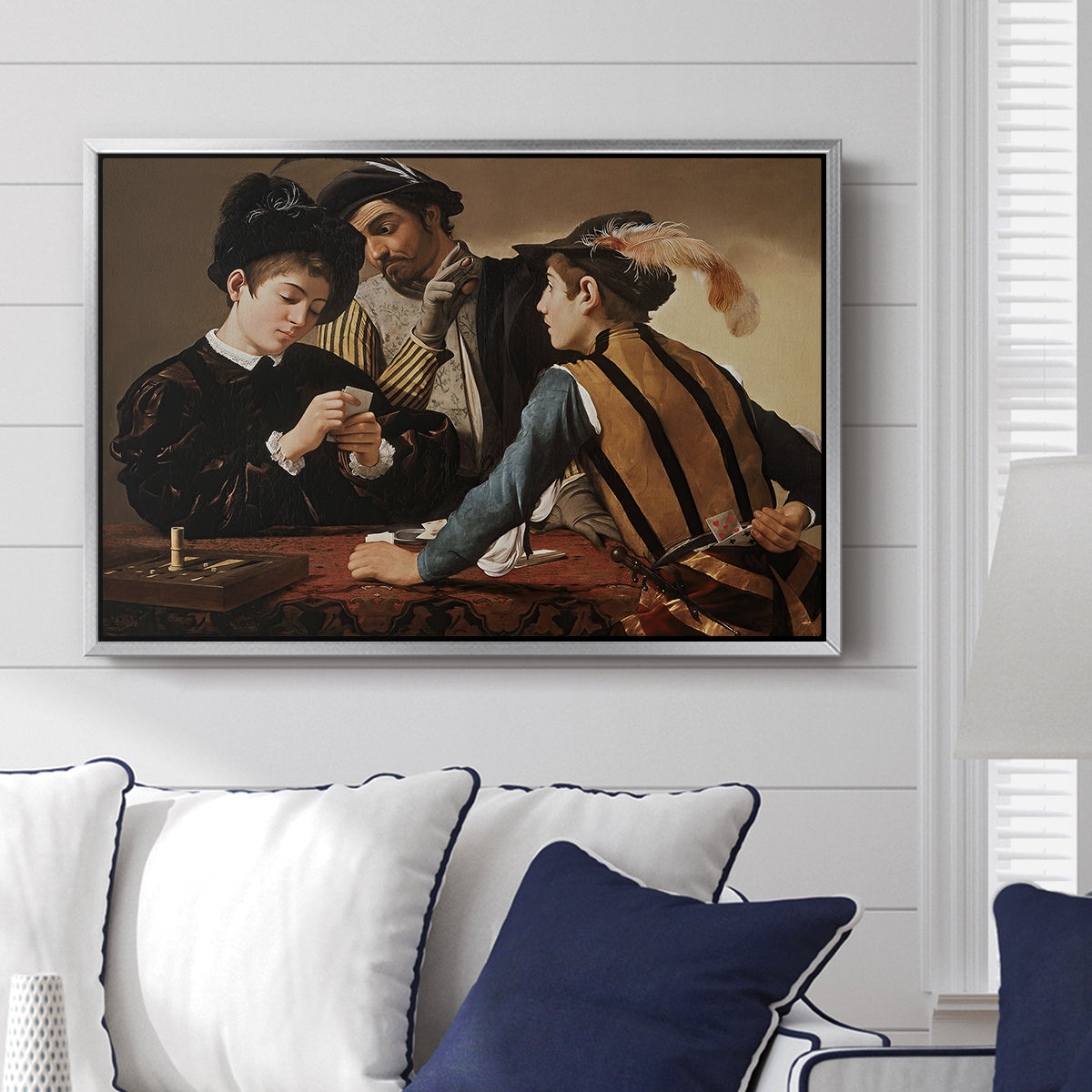 The Cardsharps, c. 1594 by Caravaggio displayed above a sofa in a living room in a silver floater frame, featuring a genre scene of card game with deception and hidden accomplice.