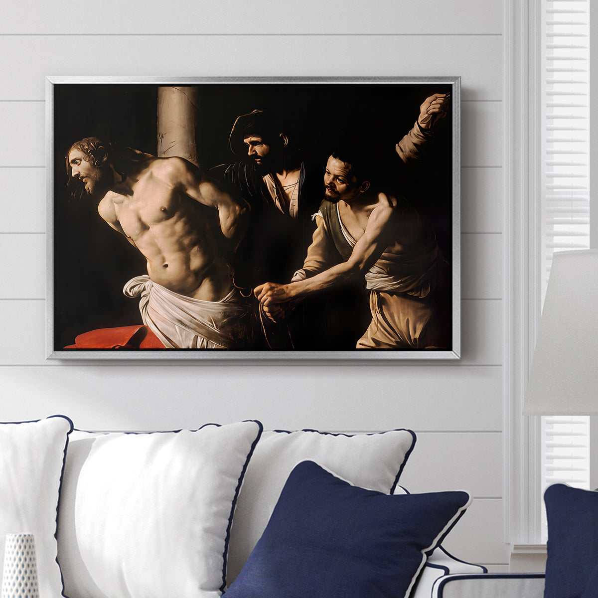 The Flagellation of Christ at the Column (Rouen Version), 1606 by Caravaggio displayed above a sofa in a living room in a silver floater frame, featuring a dramatic religious scene of Christ enduring flagellation in dark interior.