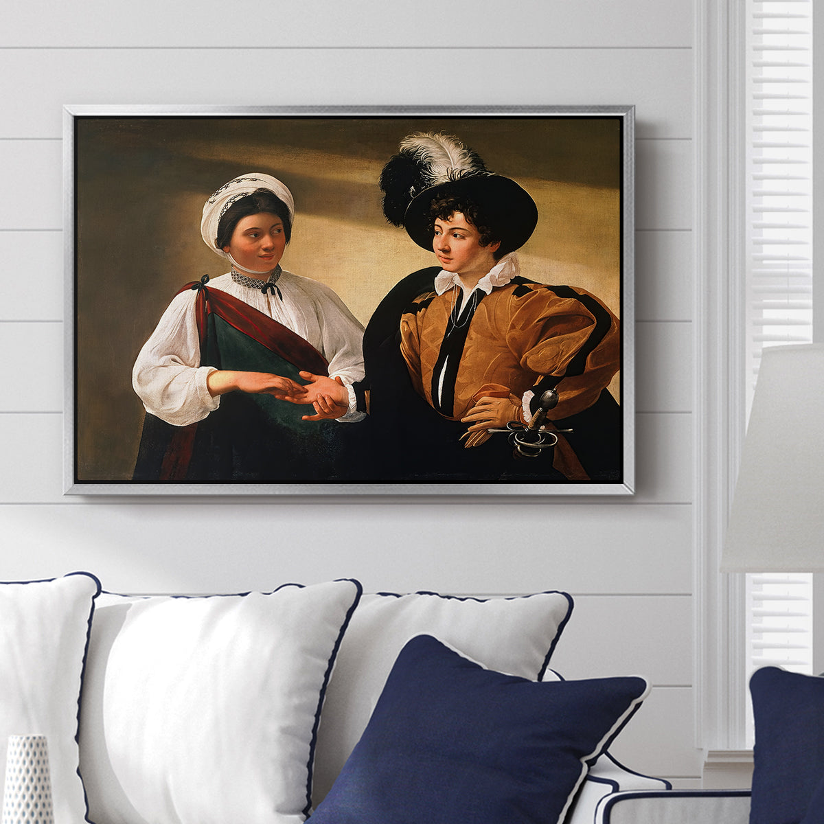 The Fortune Teller (Louvre Version), c. 1595 by Caravaggio displayed above a sofa in a living room in a silver floater frame, featuring a fortune teller examining hand of man as she slyly removes his ring.