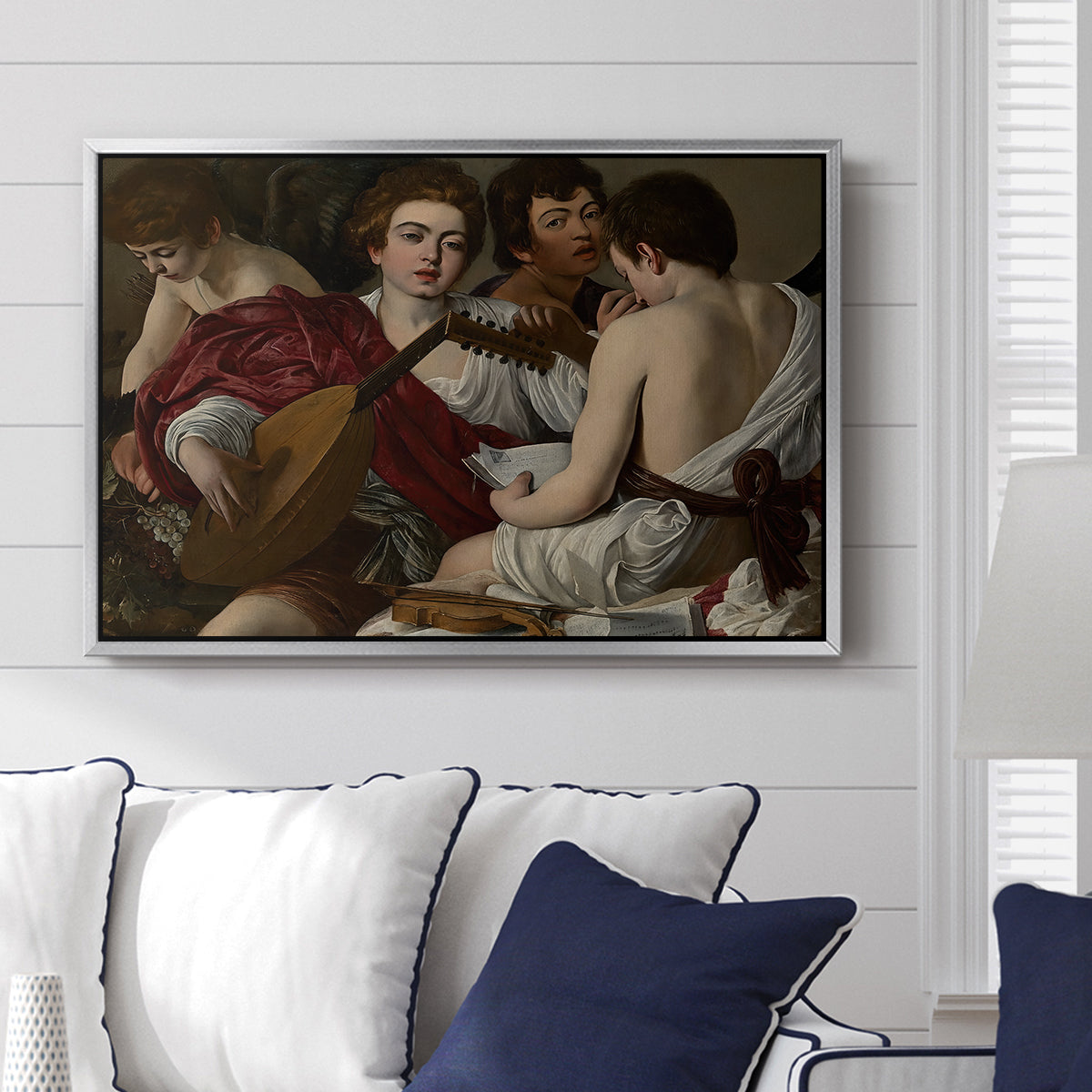 The Musicians, c. 1595 by Caravaggio displayed above a sofa in a living room in a silver floater frame, featuring an ensemble of musicians holding lute and singing in close arrangement.