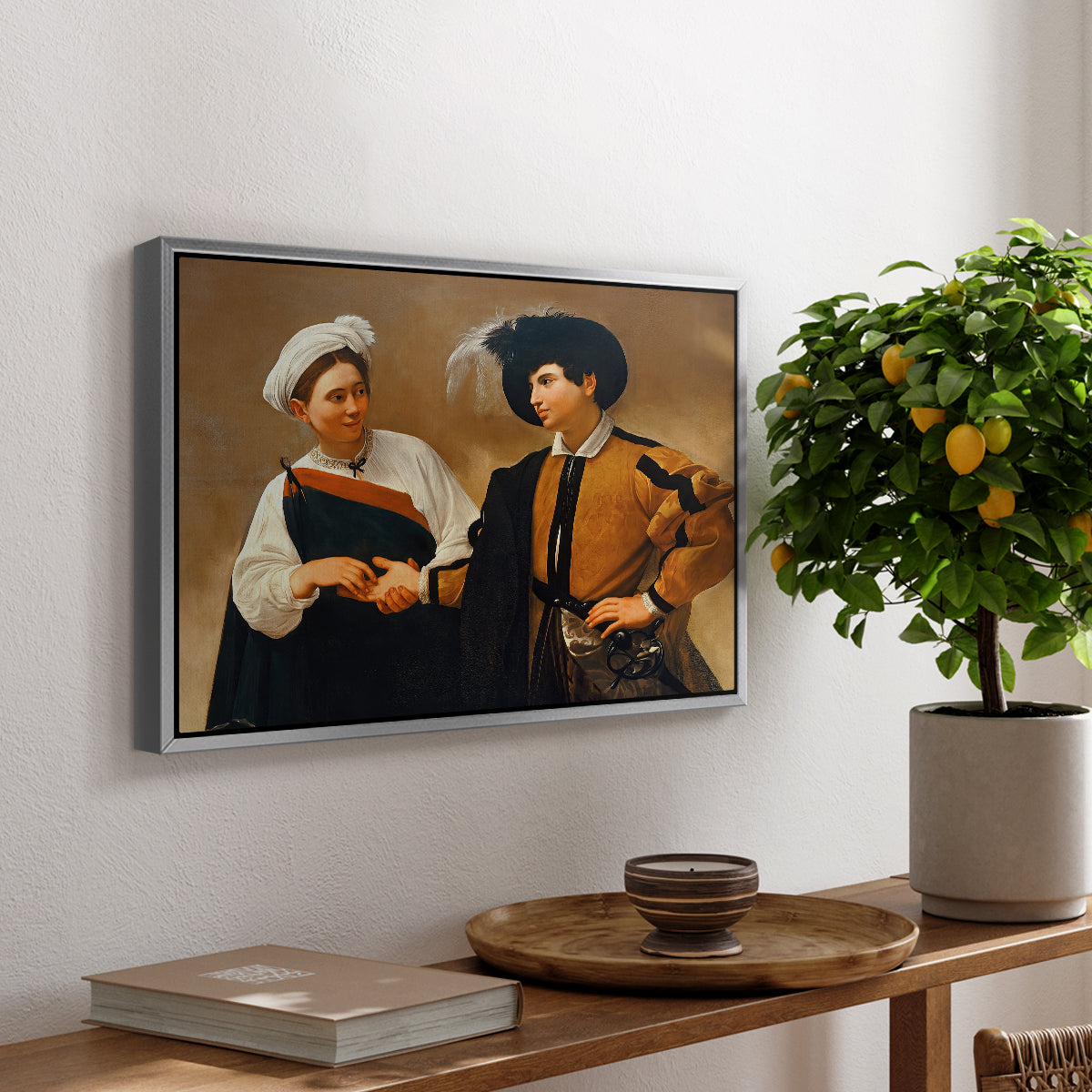 The Fortune Teller (Capitoline Version), c. 1599 by Caravaggio, presented in a silver floater framed canvas featuring a woman reading man's palm while discreetly removing his ring, displayed above a hallway console table.