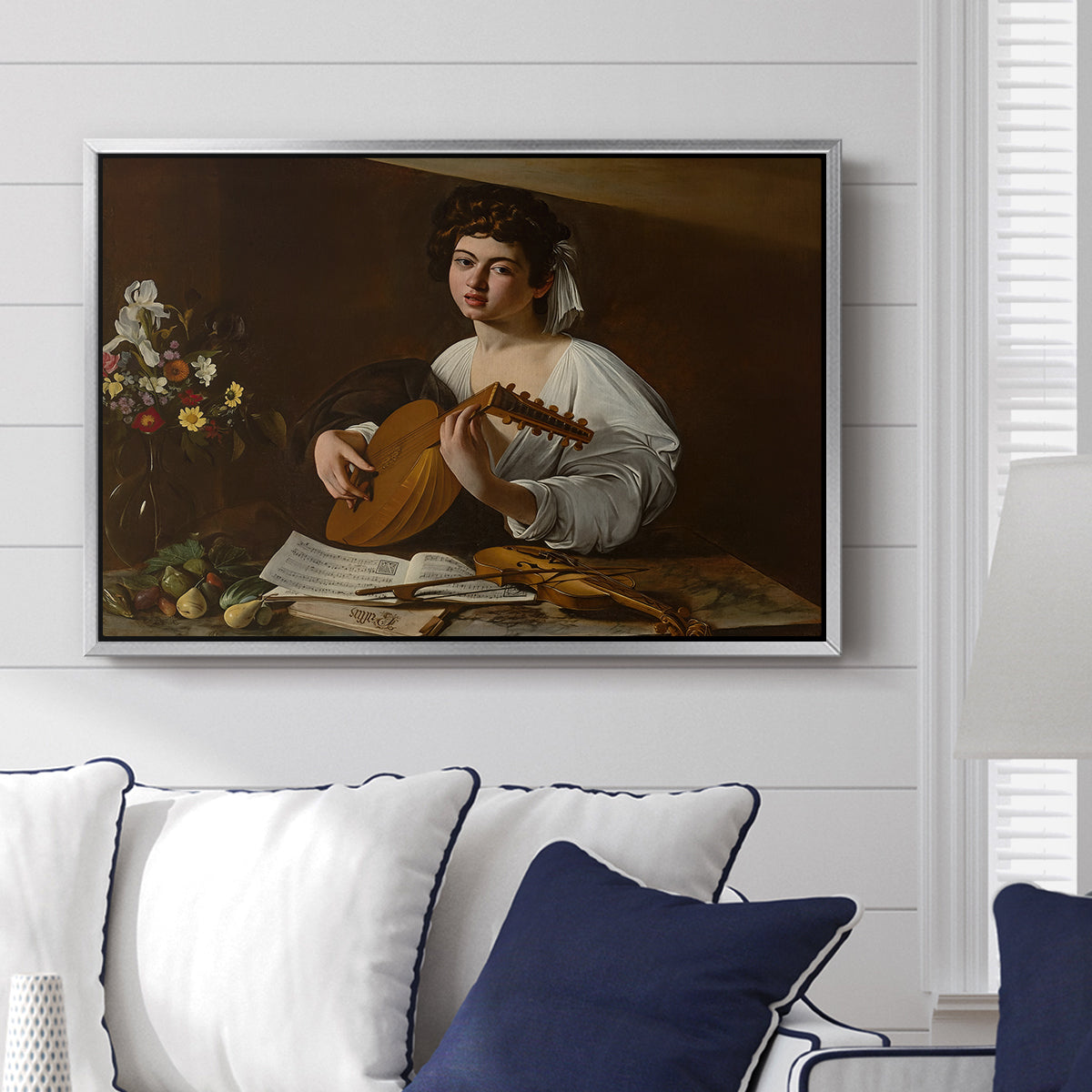 The Lute Player (Hermitage Version), c. 1596 by Caravaggio displayed above a sofa in a living room in a silver floater frame, featuring a portrait of lute player surrounded by still life arrangement.