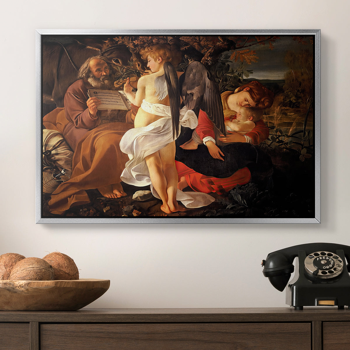 Rest on the Flight into Egypt, c. 1597 by Caravaggio in a silver floater framed canvas displayed above a console table in a hallway, featuring a Baroque landscape with angel playing music for resting travelers.
