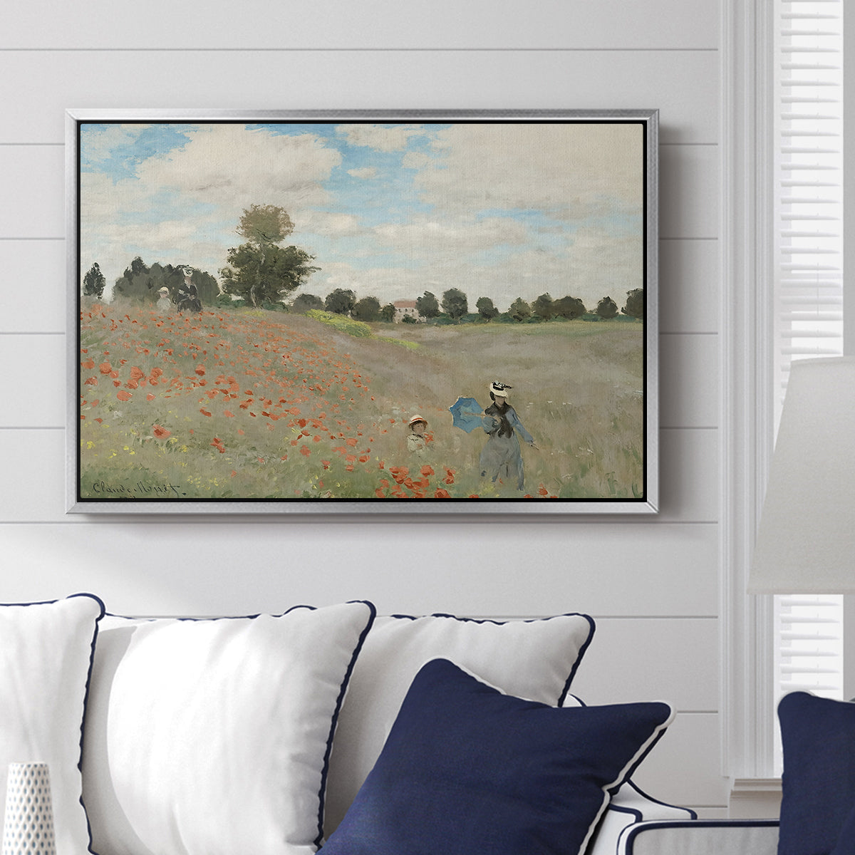 The Poppy Field near Argenteuil, 1873 by Claude Monet displayed above a sofa in a living room in a silver floater frame, featuring an impressionist countryside scene with flowers and strolling figures.