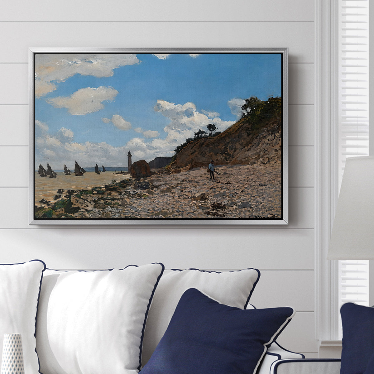 The Beach at Honfleur, 1864 by Claude Monet displayed above a sofa in a living room in a silver floater frame, featuring an impressionist seaside with people and sailboats under bright sky.