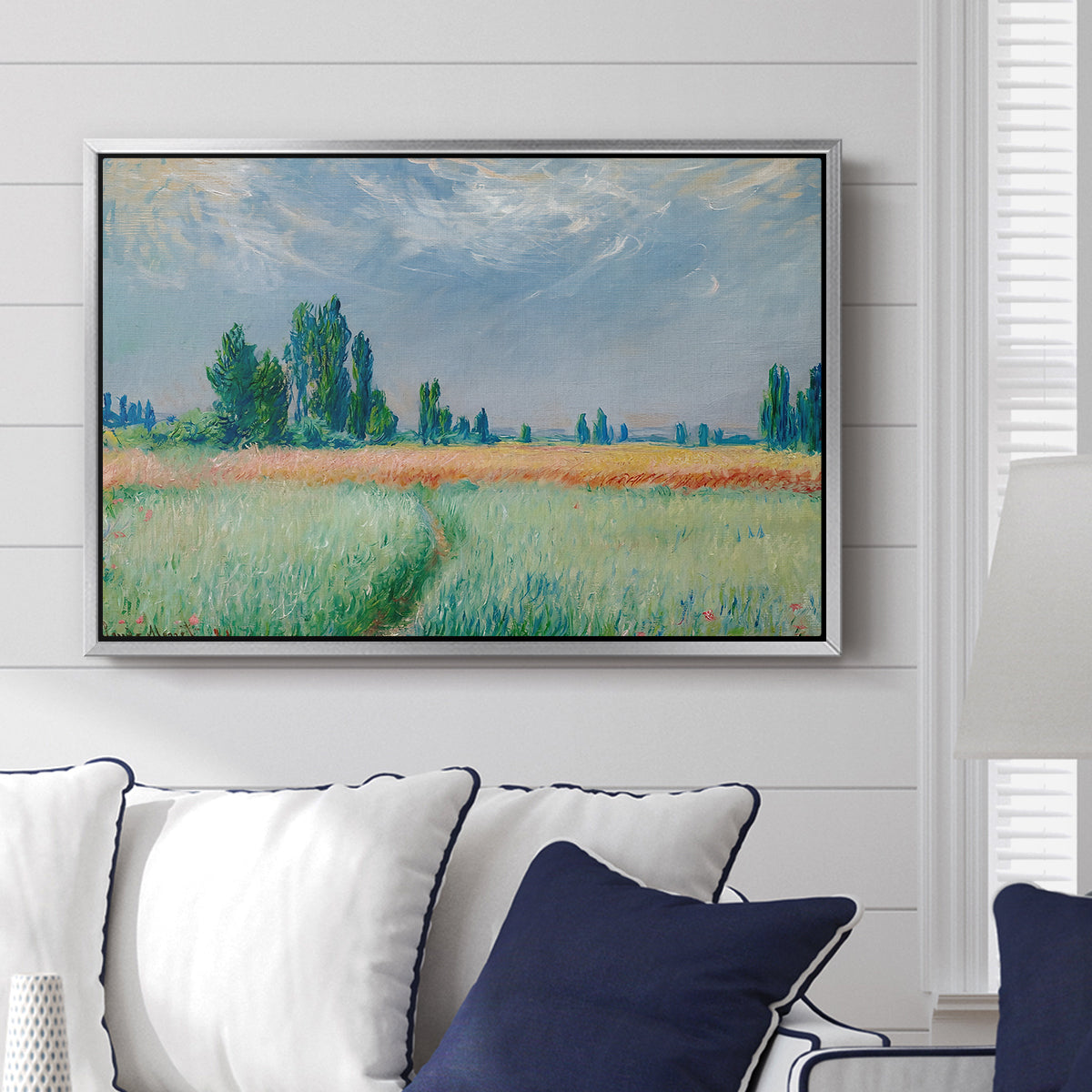 The Wheat Field (Champ de Blé), 1881 by Claude Monet displayed above a sofa in a living room in a silver floater frame, featuring an impressionist depiction of ripened grain field in countryside.