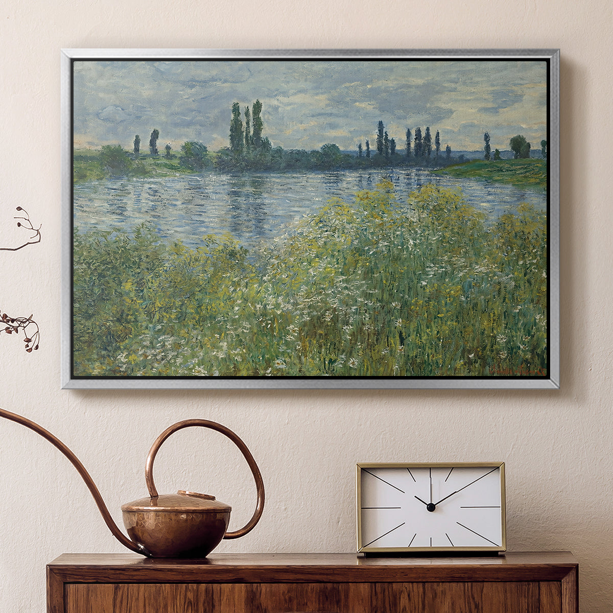 Silver floater framed canvas featuring a tranquil riverside scene with houses and vegetation, displayed above a cabinet in a soft-toned living room.