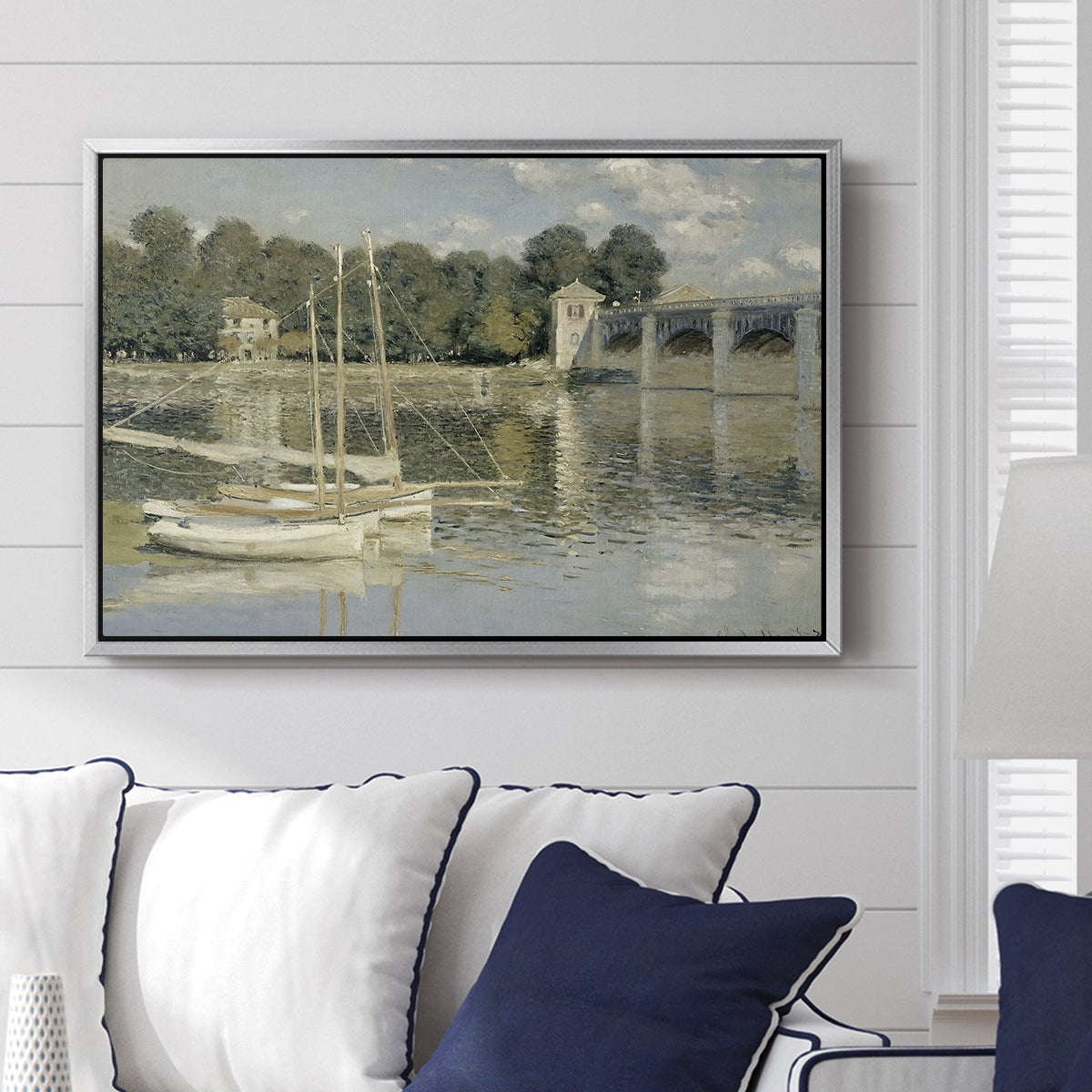 The Bridge at Argenteuil, 1874 by Claude Monet displayed above a sofa in a living room in a silver floater frame, featuring an impressionist river scene with boats passing beneath bridge.
