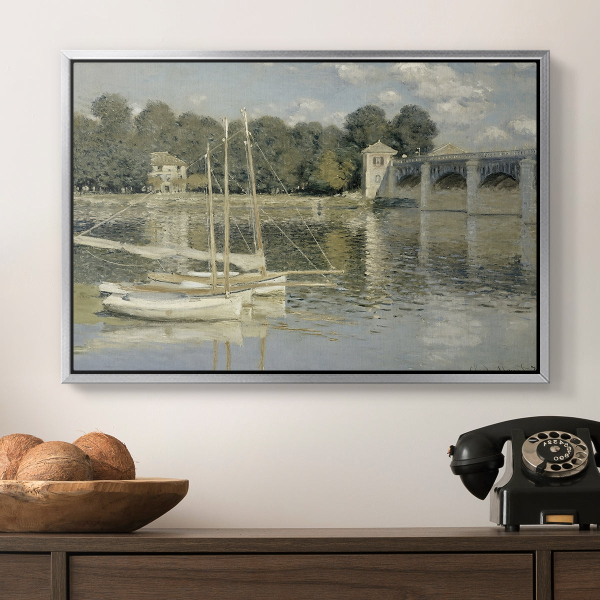 The Bridge at Argenteuil, 1874 by Claude Monet in a silver floater framed canvas displayed above a console table in a hallway, featuring a riverside view with architecture and nautical activity.