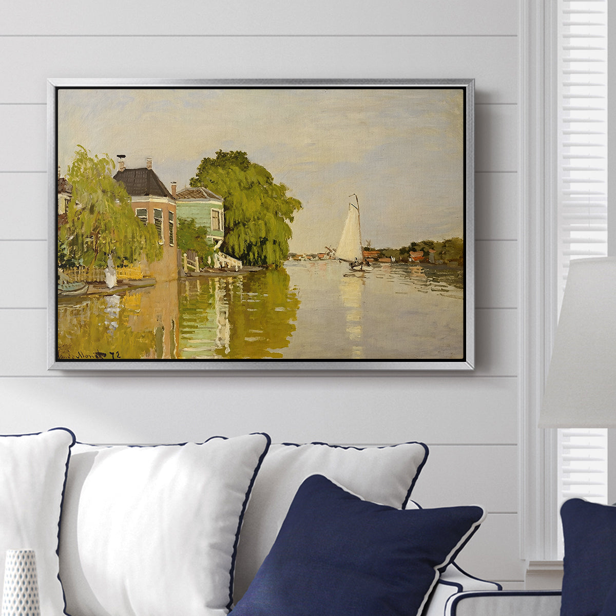 Houses on the Achterzaan, 1871 by Claude Monet displayed above a sofa in a living room in a silver floater frame, featuring riverside homes with trees and mirrored reflections in water.