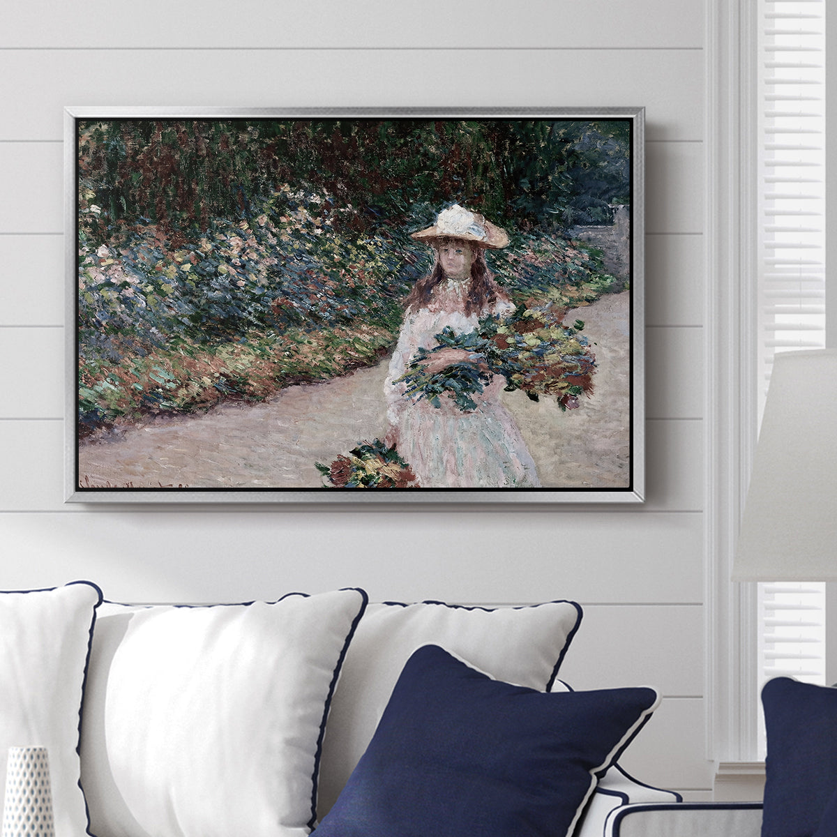 Young Girl in the Garden at Giverny, 1888 by Claude Monet displayed above a sofa in a living room in a silver floater frame, featuring an impressionist garden scene with child among greenery and blossoms.