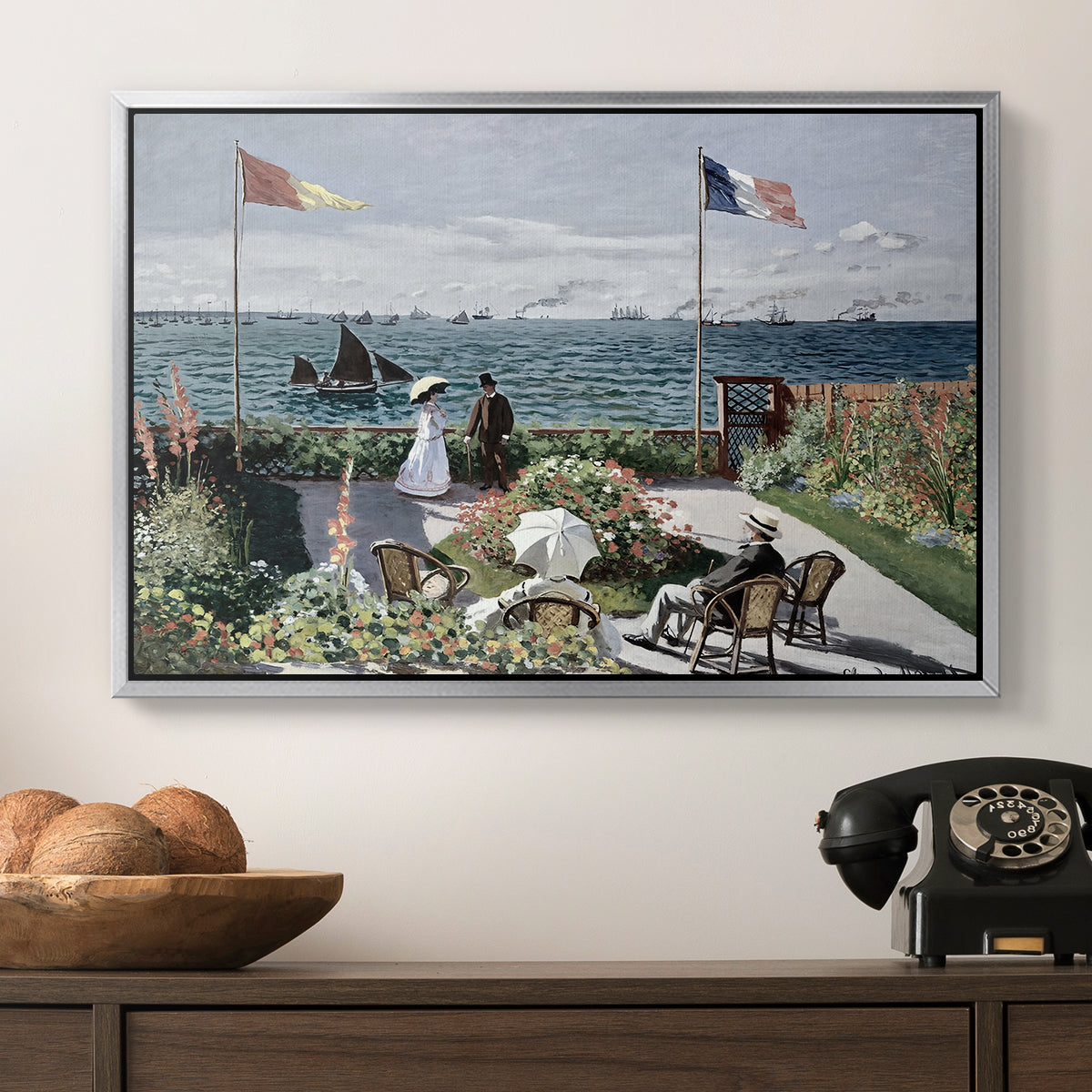 Garden at Sainte-Adresse, 1867 by Claude Monet in a silver floater framed canvas displayed above a console table in a hallway, featuring a landscaped terrace with figures and flags near shoreline.