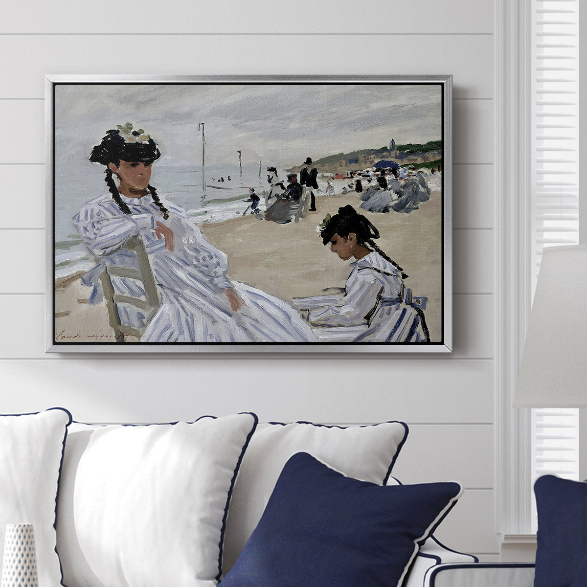 On the Beach at Trouville, 1870 by Claude Monet displayed above a sofa in a living room in a silver floater frame, featuring an impressionist seaside scene with women in dresses and hats.