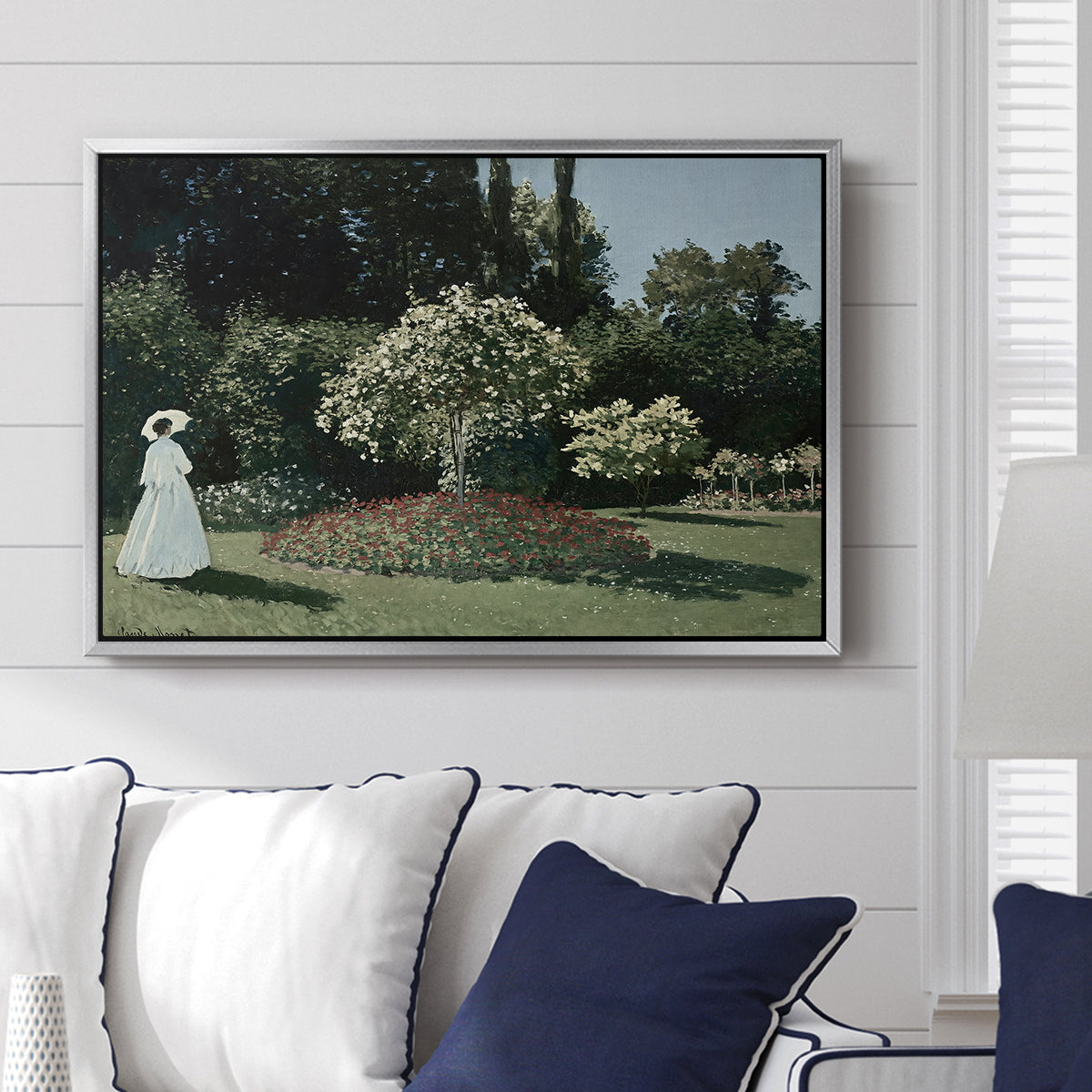 Woman in the Garden, Sainte-Adresse, 1867 by Claude Monet displayed above a sofa in a living room in a silver floater frame, featuring an impressionist figure in sunlit garden with lush vegetation.