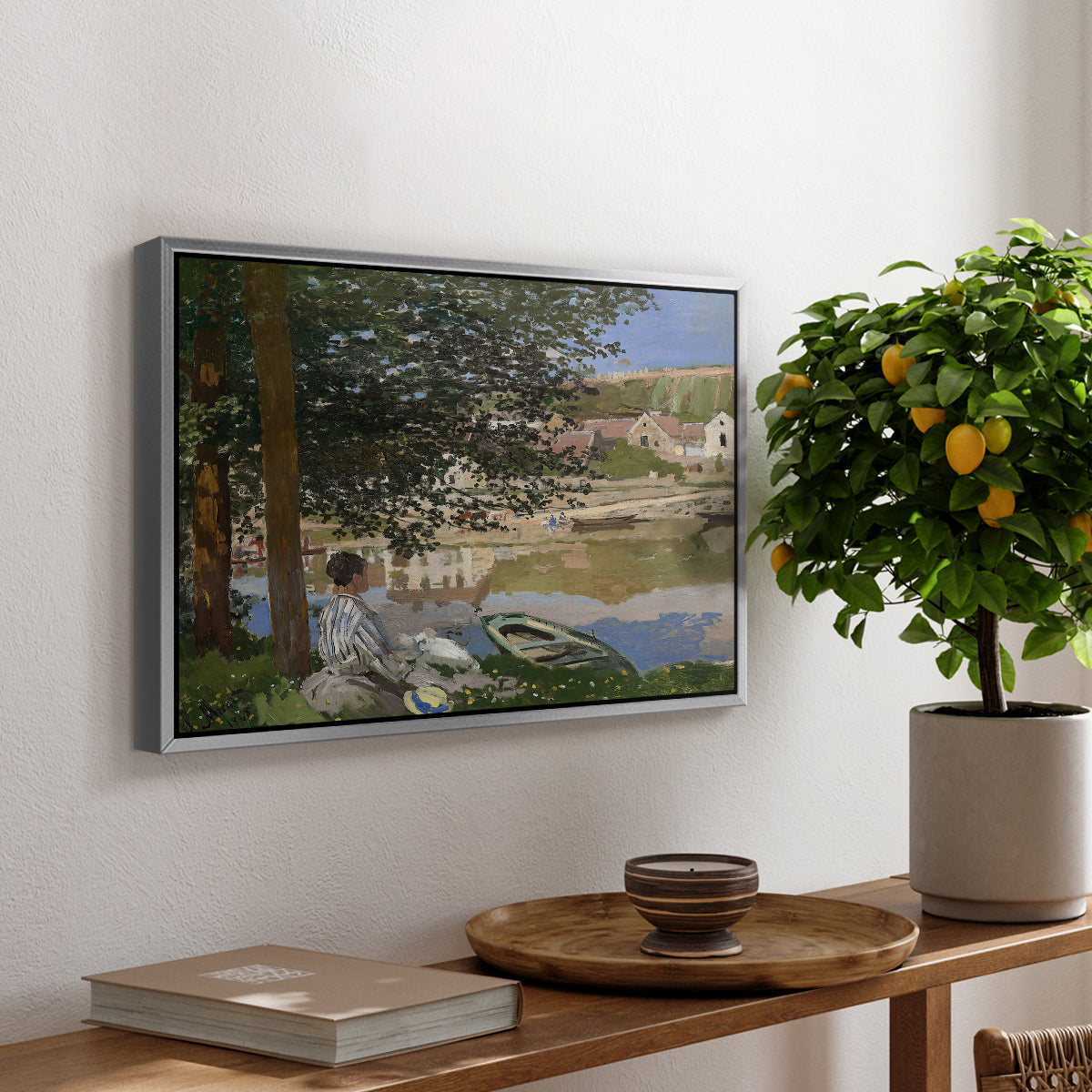 On the Bank of the Seine, Bennecourt, 1868 by Claude Monet, presented in a silver floater framed canvas featuring a woman and child seated along riverbank beneath trees in soft daylight, displayed above a hallway console table.