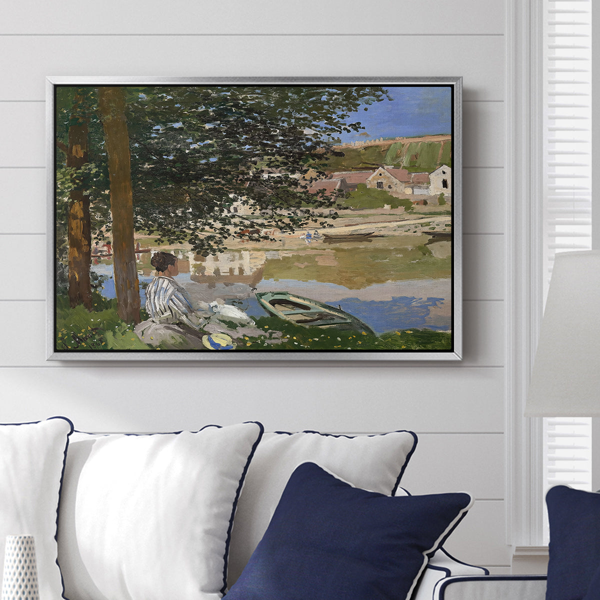 On the Bank of the Seine, Bennecourt, 1868 by Claude Monet displayed above a sofa in a living room in a silver floater frame, featuring a peaceful riverside scene with figures resting in grassy area by water.