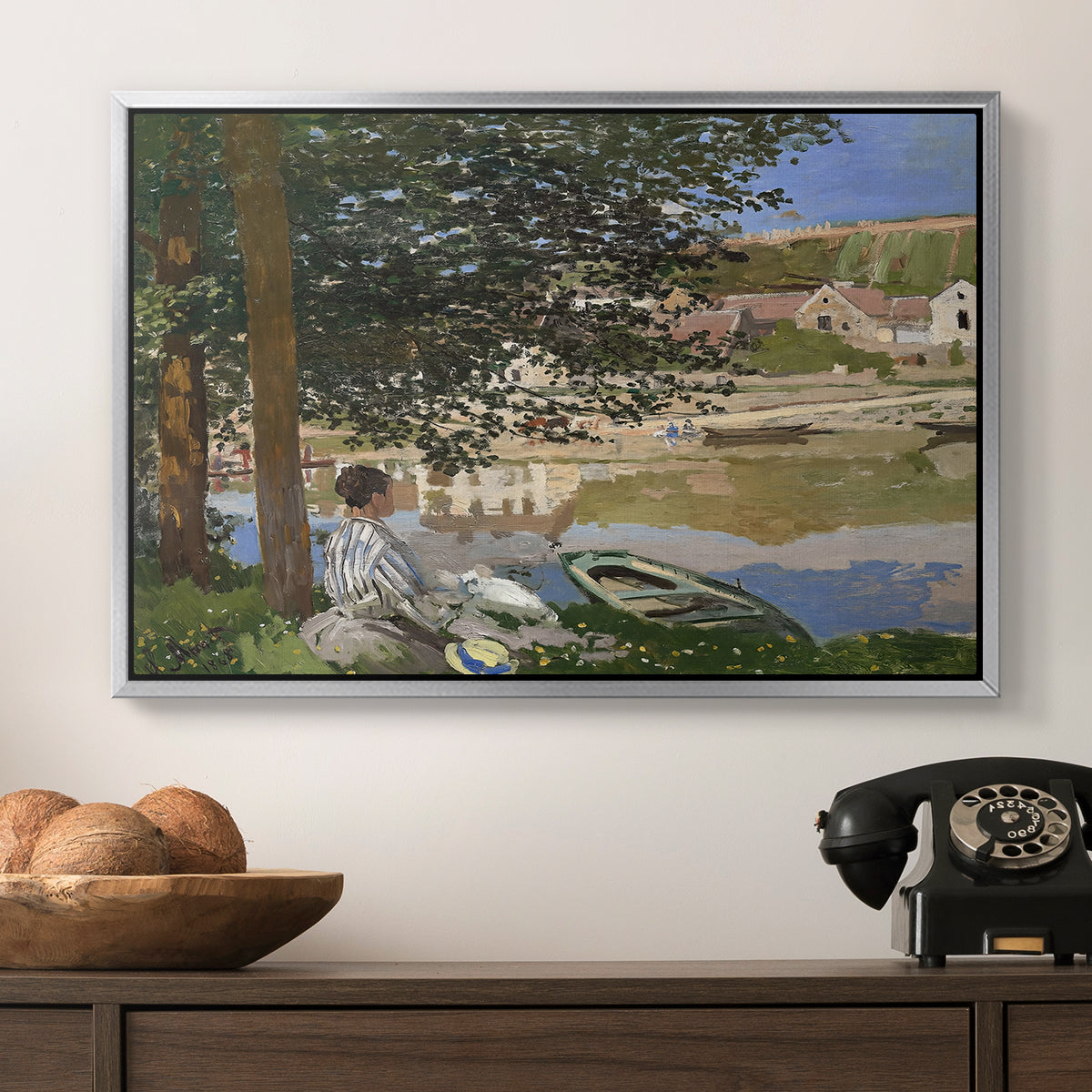 On the Bank of the Seine, Bennecourt, 1868 by Claude Monet in a silver floater framed canvas displayed above a console table in a hallway, featuring a quiet outdoor setting with figures near river under foliage.