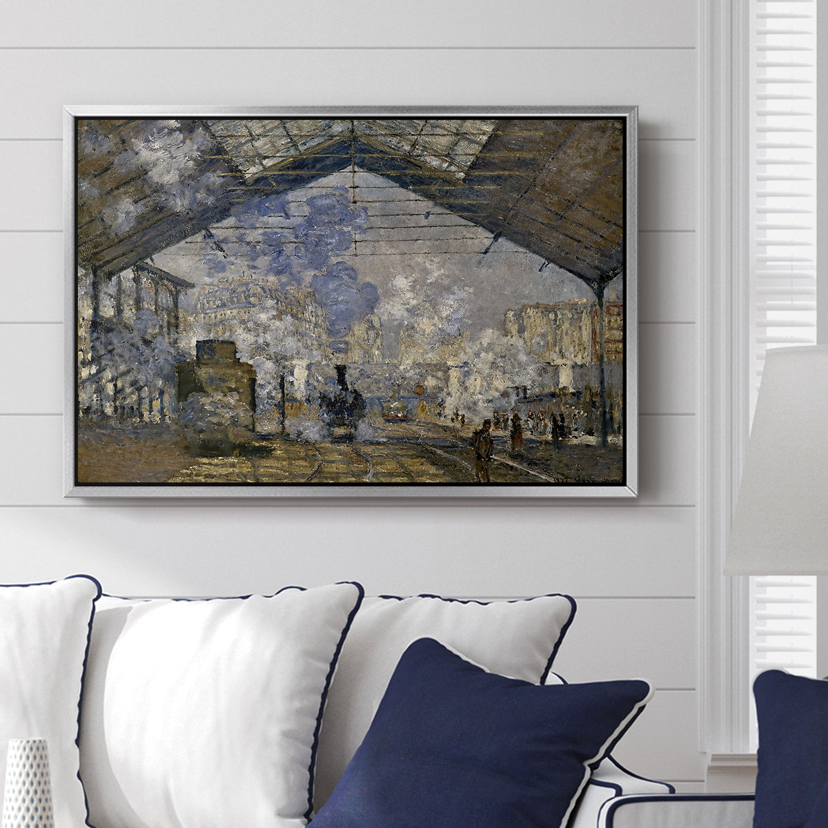 The Gare Saint-Lazare, 1877 by Claude Monet displayed above a sofa in a living room in a silver floater frame, featuring an impressionist scene of trains and smoke inside busy railway station.