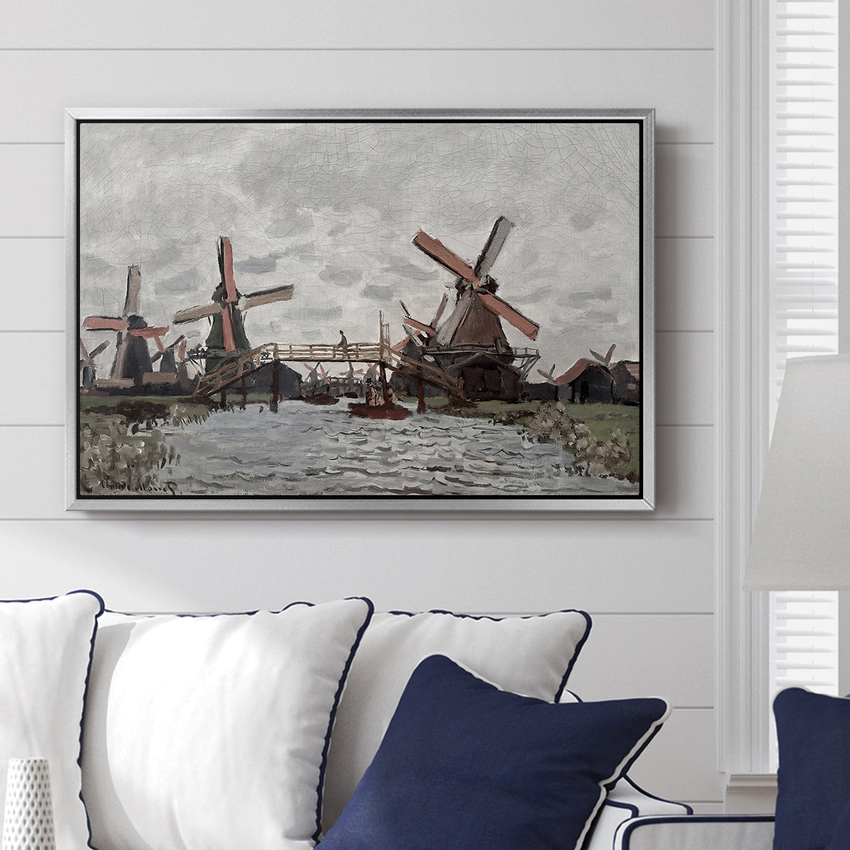 Windmills near Zaandam, 1871 by Claude Monet displayed above a sofa in a living room in a silver floater frame, featuring an impressionist view of Dutch windmills along riverbank.