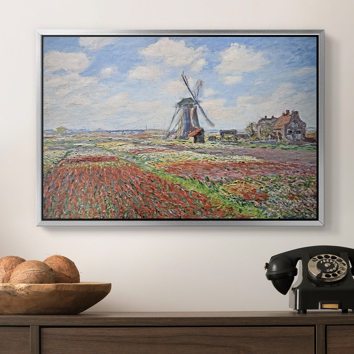 Tulip Fields with the Rijnsburg Windmill, 1886 by Claude Monet in a silver floater framed canvas displayed above a console table in a hallway, featuring a rural scene with patterned rows of flowers and windmill.