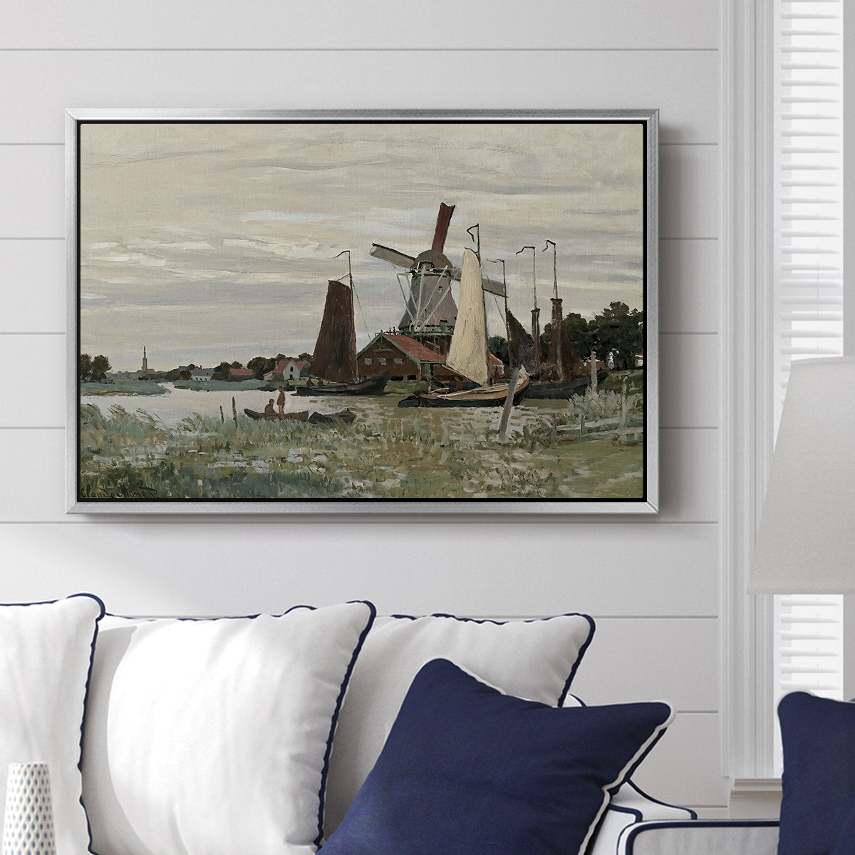 Windmill at Zaandam, 1871 by Claude Monet displayed above a sofa in a living room in a silver floater frame, featuring an impressionist depiction of windmill near riverbank.