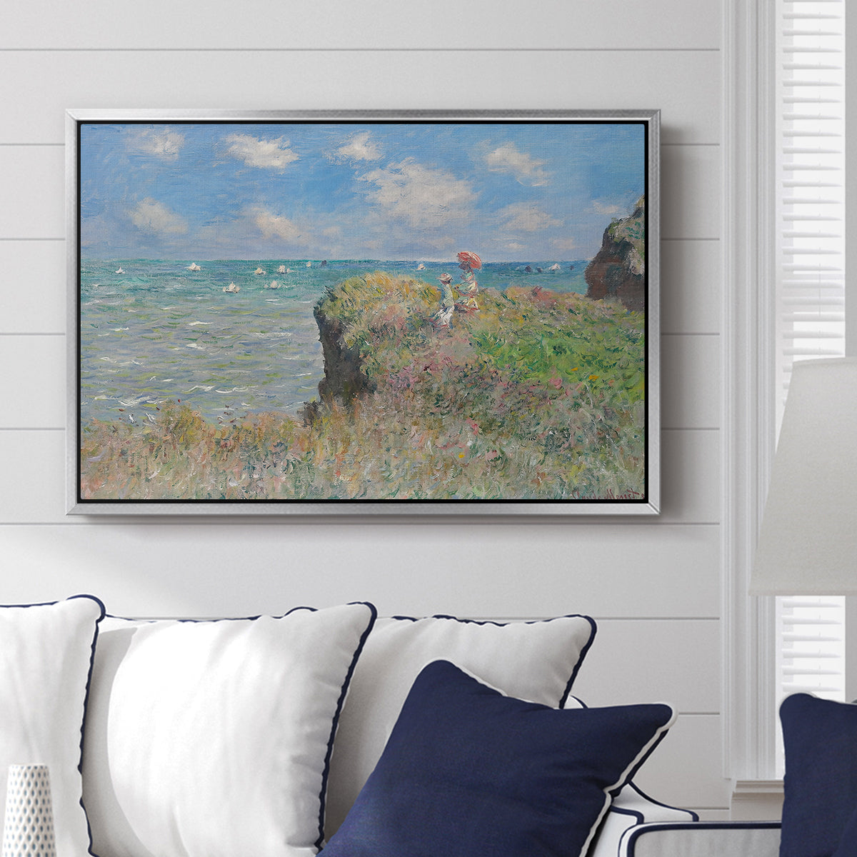 The Cliff Walk at Pourville, 1882 by Claude Monet displayed above a sofa in a living room in a silver floater frame, featuring an impressionist seaside landscape with people strolling above water.