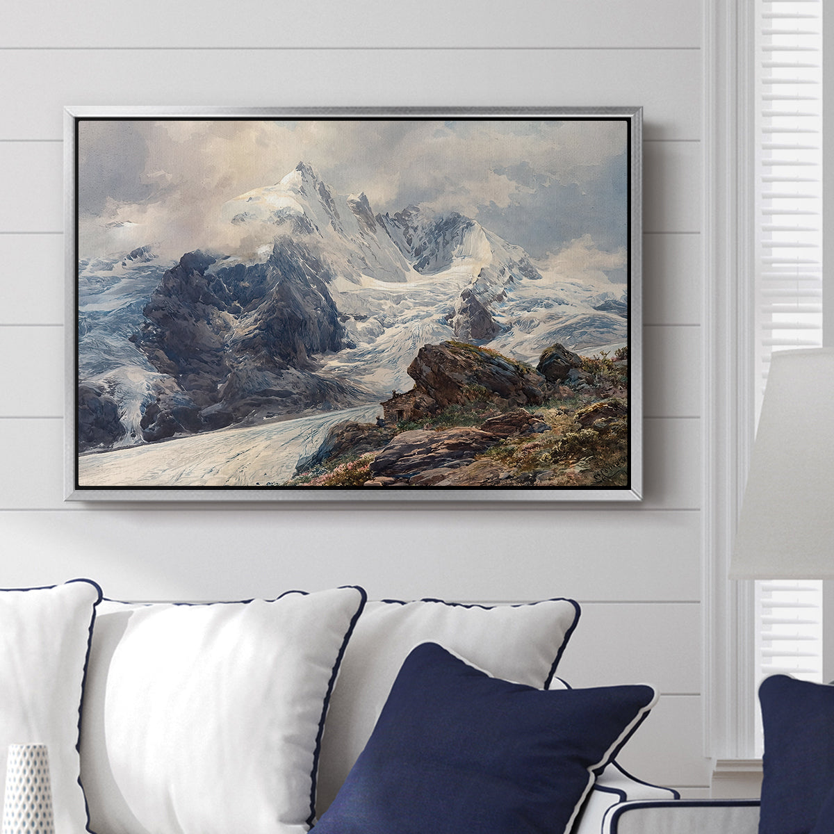 The Grossglockner, 1880 by Edward Theodore Compton displayed above a sofa in a living room in a silver floater frame, featuring a dramatic mountain summit with snow and rugged slopes.