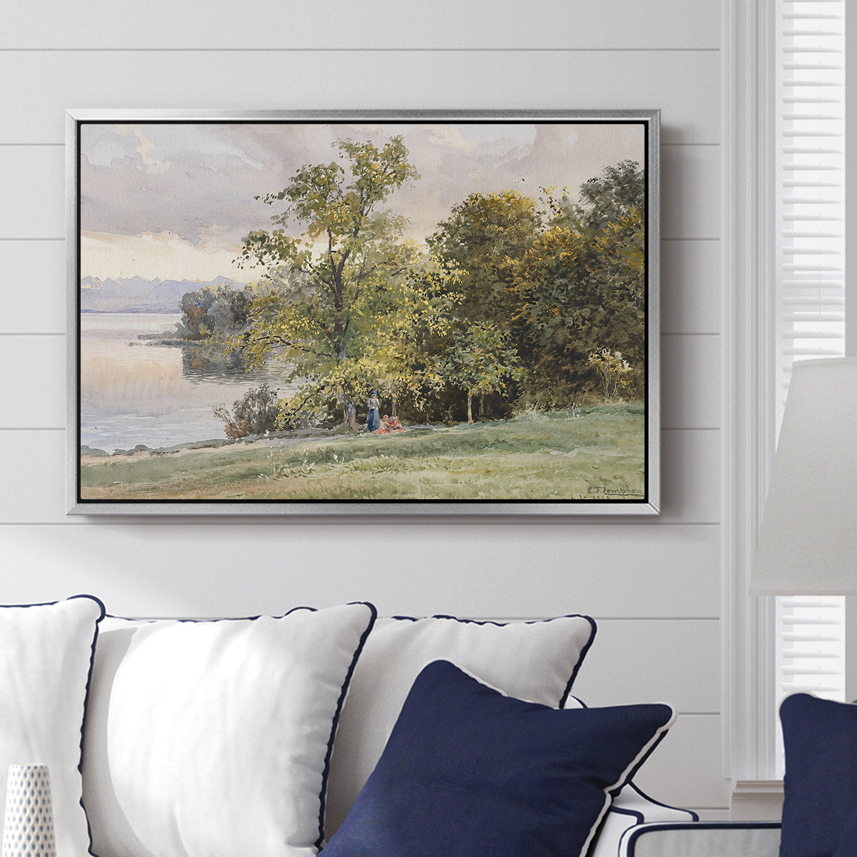 Landscape at Lake Starnberg, 1890 by Edward Theodore Compton displayed above a sofa in a living room in a silver floater frame, featuring a peaceful lake scene with reflections and surrounding greenery.